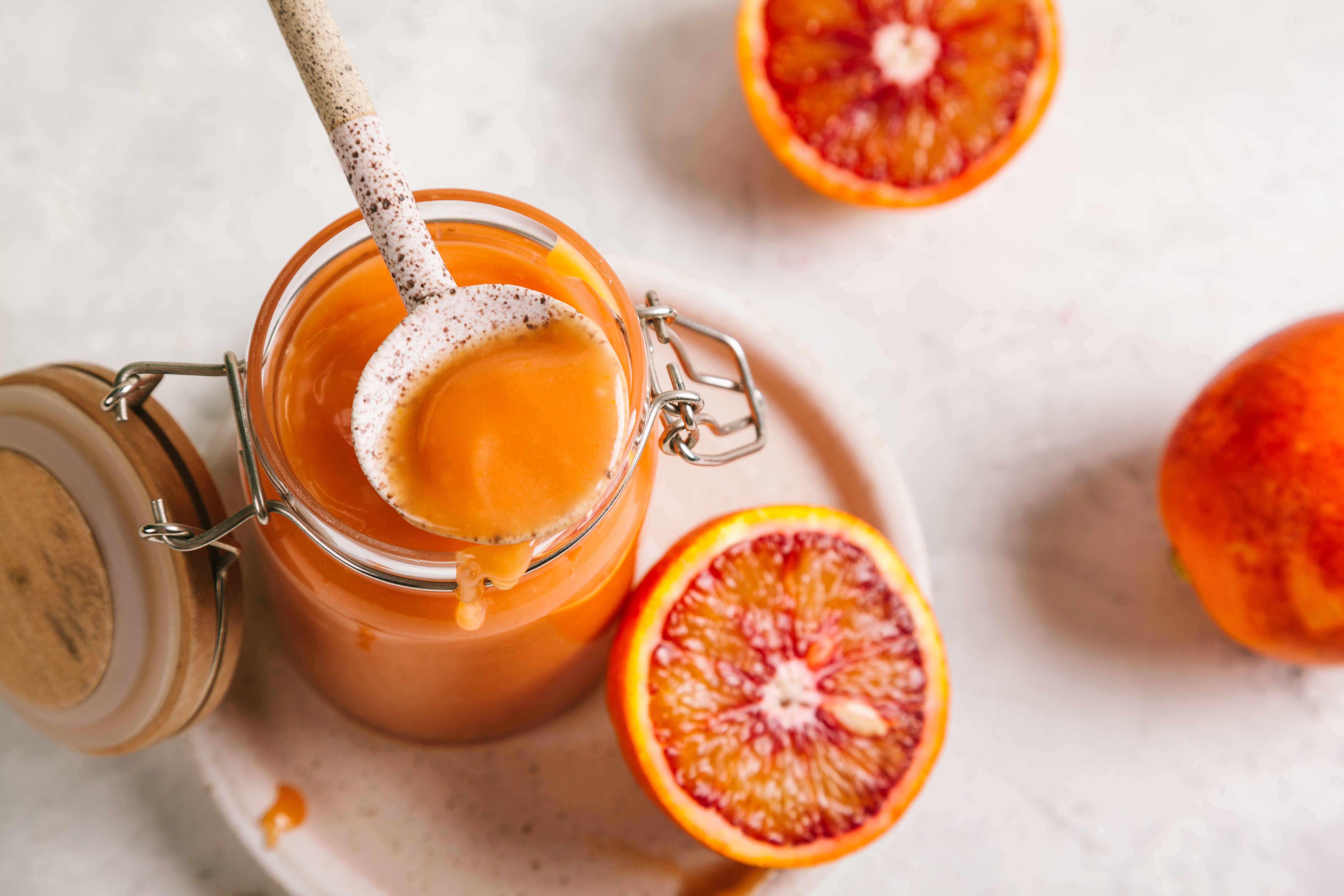 Jar of smooth blood orange curd with a spoonful lifted above the surface, surrounded by halved blood oranges on a light background.